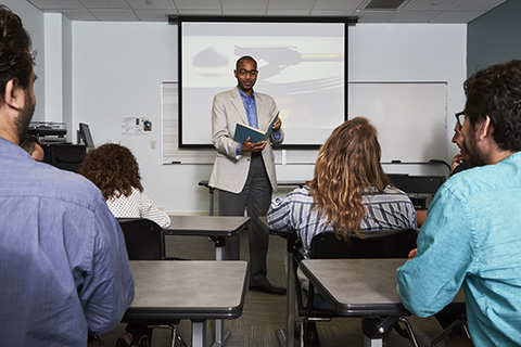 A professor stands at the front of a class with an open book